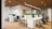 A modern office space with an open-plan layout, featuring multiple white desks arranged in a row and equipped with sleek computers. Comfortable office chairs are positioned around the desks. Green plants are placed on the partitioned tops of the desks, adding a touch of nature to the environment. The room has wooden flooring and a ceiling with exposed wooden beams, along with contemporary lighting fixtures. Artworks hang on the walls, contributing to a creative atmosphere.