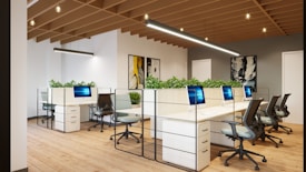 A modern office space with an open-plan layout, featuring multiple white desks arranged in a row and equipped with sleek computers. Comfortable office chairs are positioned around the desks. Green plants are placed on the partitioned tops of the desks, adding a touch of nature to the environment. The room has wooden flooring and a ceiling with exposed wooden beams, along with contemporary lighting fixtures. Artworks hang on the walls, contributing to a creative atmosphere.