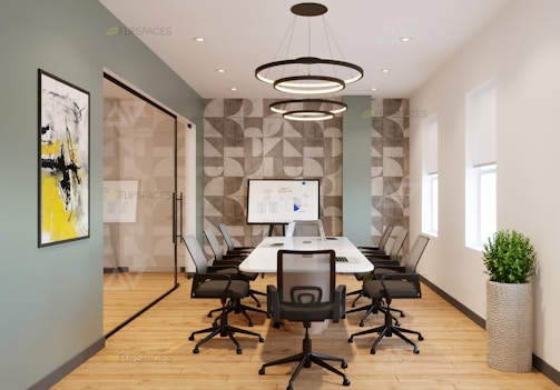 A modern conference room with a long table surrounded by black office chairs. A large piece of abstract art with yellow and black accents is on the wall. The floor is wooden, and there is a potted plant in the corner. The walls feature geometric patterns, and a whiteboard with a graph is visible at the far end of the room. Circular overhead lights provide illumination.