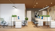 An open-plan office space features a modern, minimalist design with several workstations arranged in rows. Each workstation is equipped with a computer and ergonomic chair. Green plants are placed between sections to add a natural element. The room has wooden flooring, and a geometric patterned wall serves as an accent. Pendant lighting fixtures hang from the wooden slatted ceiling.