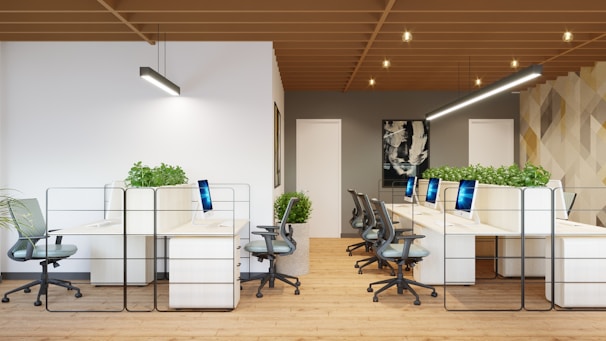 An open-plan office space features a modern, minimalist design with several workstations arranged in rows. Each workstation is equipped with a computer and ergonomic chair. Green plants are placed between sections to add a natural element. The room has wooden flooring, and a geometric patterned wall serves as an accent. Pendant lighting fixtures hang from the wooden slatted ceiling.
