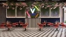 A modern lounge interior featuring geometric floor tiles, pink chairs with gold legs, and wooden tables. The back wall is adorned with vertical neon lights and lush greenery, including pink flowers. Disco balls hang from the ceiling, adding a festive touch.