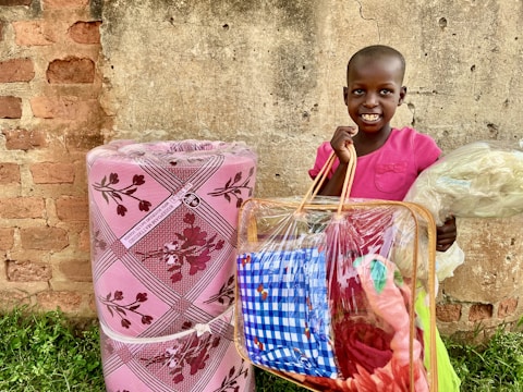 A child is smiling and holding a clear plastic bag filled with colorful items, including a blue and white patterned cloth, while standing in front of a textured brick and concrete wall. Next to the child is a large rolled-up foam mattress wrapped in pink plastic with floral designs.