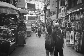 A busy marketplace corridor filled with stalls displaying various clothing and items. The walkway is lined with shoppers moving through, some carrying backpacks and bags. Overhead signage is visible along with arches and hanging decorations that suggest an indoor market setting.