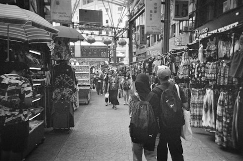 A busy marketplace corridor filled with stalls displaying various clothing and items. The walkway is lined with shoppers moving through, some carrying backpacks and bags. Overhead signage is visible along with arches and hanging decorations that suggest an indoor market setting.