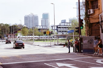 A city street scene featuring multiple cars at a traffic light. Several pedestrians are waiting to cross the street, standing near a bus stop and a street sign that reads 'Station MRT Pasar Seni'. The background includes tall modern buildings and trees lining the roadside. The atmosphere is urban with a mixture of greenery and architecture.