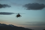 A helicopter is flying against the backdrop of a scenic mountain range at dusk. The sky is filled with soft hues of blue, orange, and pink, with a few scattered clouds. The silhouette of the helicopter is highlighted against the sky, adding a sense of motion and adventure.