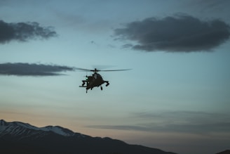 A helicopter is flying against the backdrop of a scenic mountain range at dusk. The sky is filled with soft hues of blue, orange, and pink, with a few scattered clouds. The silhouette of the helicopter is highlighted against the sky, adding a sense of motion and adventure.
