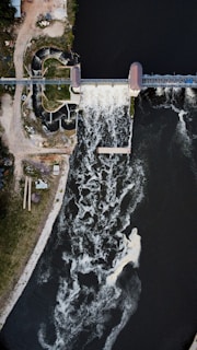 An aerial view of a dam with water flowing over it, creating a frothy pattern in the river below. The surrounding area includes grassy land on one side and structures near the dam. The flow of water is dynamic and prominent, with some white rapids visible.