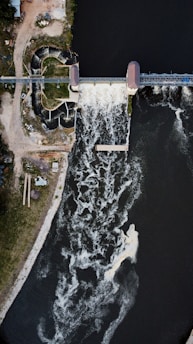 An aerial view of a dam with water flowing over it, creating a frothy pattern in the river below. The surrounding area includes grassy land on one side and structures near the dam. The flow of water is dynamic and prominent, with some white rapids visible.
