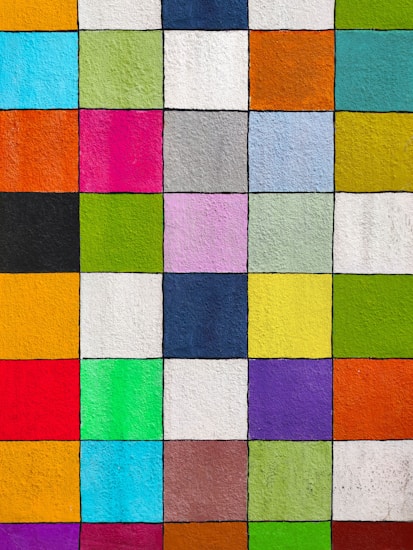 A vibrant grid of colorful squares, each filled with a different bold hue. The squares are evenly divided, creating a patchwork of colors including shades of red, blue, green, yellow, pink, purple, and orange. The texture of the surface is slightly rough, suggesting a painted wall or street art canvas.