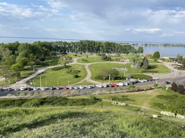 A scenic park view features lush greenery, a lake in the background, and well-maintained lawns. Pathways wind through the park, with people walking and relaxing. A line of parked cars is visible along the road bordering the park.