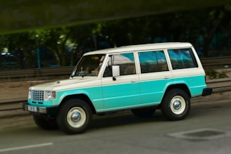 A vintage SUV with two-tone coloring featuring a teal and white paint job, traveling on a road. The background is slightly blurred, suggesting motion, with trees and a metal barrier visible.