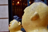 Close-up view of two large, yellow sculptures with textured surfaces, shaped like heads with stylized facial features. The foreground sculpture shows more detail with visible facial contours and an earthy texture, while the background sculpture appears slightly blurred, emphasizing depth. Both sculptures face to the left and are framed by an urban setting with blurred lights in the background.