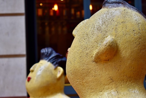 Close-up view of two large, yellow sculptures with textured surfaces, shaped like heads with stylized facial features. The foreground sculpture shows more detail with visible facial contours and an earthy texture, while the background sculpture appears slightly blurred, emphasizing depth. Both sculptures face to the left and are framed by an urban setting with blurred lights in the background.