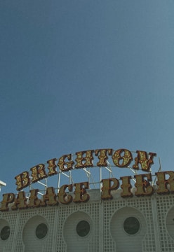 A vintage-style sign displaying the words 'BRIGHTON PALACE PIER' against a clear blue sky. The sign features retro light bulb lettering and is situated on top of a white, lattice-patterned building façade with circular windows.