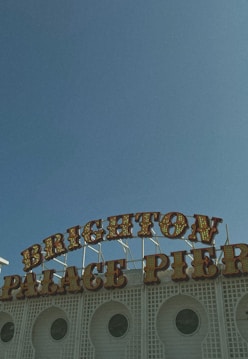 A vintage-style sign displaying the words 'BRIGHTON PALACE PIER' against a clear blue sky. The sign features retro light bulb lettering and is situated on top of a white, lattice-patterned building fa&ccedil;ade with circular windows.