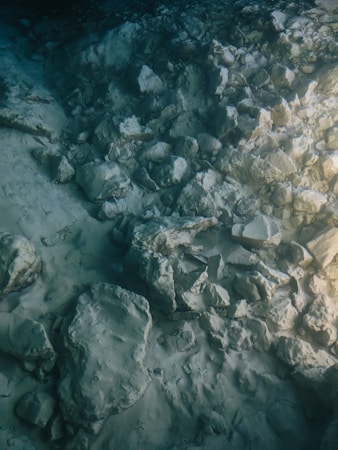 A serene underwater landscape featuring a scattering of rocks and stones on a sandy seabed. The lighting is subtle, creating a tranquil and slightly mysterious ambiance. The water's hue adds a gentle blue-green tint to the scene, enhancing the feeling of being submerged.