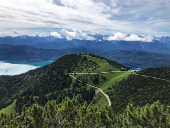 A breathtaking view of a lush green mountainous landscape with winding paths leading to the summit. In the distance, a serene turquoise lake contrasts with the dark, forested slopes. The backdrop features towering mountain ranges partially obscured by wispy clouds under an expansive sky.