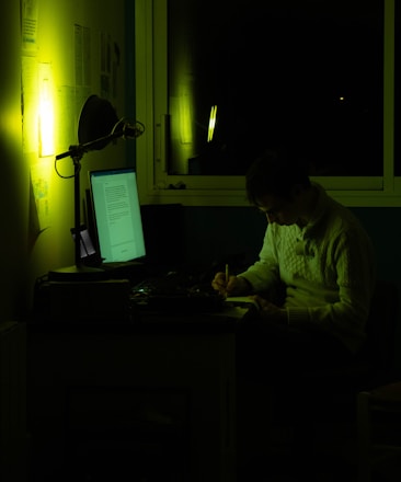 A person sits at a desk in a dimly lit room, writing on a notepad. The scene is illuminated by the greenish glow of a computer screen and a soft light on the wall. Papers are pinned to the wall and the room appears quiet and focused.