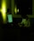 A person sits at a desk in a dimly lit room, writing on a notepad. The scene is illuminated by the greenish glow of a computer screen and a soft light on the wall. Papers are pinned to the wall and the room appears quiet and focused.