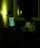 A person sits at a desk in a dimly lit room, writing on a notepad. The scene is illuminated by the greenish glow of a computer screen and a soft light on the wall. Papers are pinned to the wall and the room appears quiet and focused.