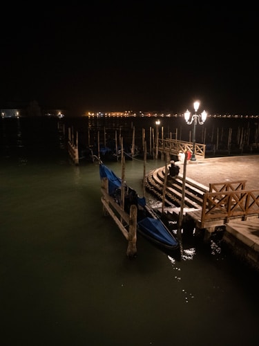 A dimly lit scene by a waterway featuring a wooden dock and gondolas tethered at the edge. A lamp post illuminates the dock area with a warm glow, casting reflections on the water. The background shows a row of distant lights along the water's edge, possibly indicating buildings or a waterfront promenade.