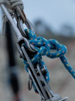 A close-up view of a metal climbing gear setup, featuring a carabiner linked with a strong blue climbing rope. The rope has orange and green speckles and is securely tied around the gear. The image is sharp, with the focus on the metal and rope, while the background is blurred, suggesting an outdoor environment.