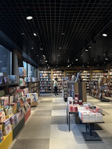 A well-lit bookstore interior featuring multiple shelves filled with books. The floor is tiled and there are various categories of books displayed on both sides. A person stands in the background, browsing through the collection. The ceiling has a grid pattern with spotlights providing illumination.
