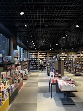 A well-lit bookstore interior featuring multiple shelves filled with books. The floor is tiled and there are various categories of books displayed on both sides. A person stands in the background, browsing through the collection. The ceiling has a grid pattern with spotlights providing illumination.