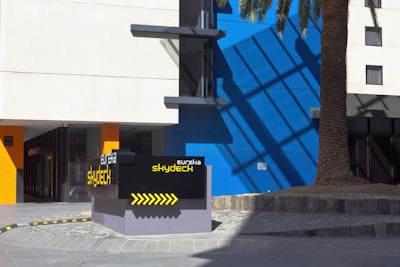 A modern building facade with a prominent sign for 'Eureka Skydeck' in front. There's a large palm tree casting a shadow on the blue and white exterior wall. The architecture features sleek lines and bold colors with a combination of blue, white, and orange.
