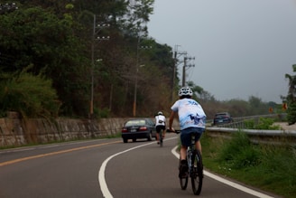 Two cyclists ride along a curving road surrounded by greenery. One cyclist wears a helmet and a white shirt with graphics on the back. The road is bordered by a metal guardrail on one side and a concrete wall on the other. Several vehicles are visible on the road, including a black car following the cyclists.