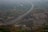 A long highway stretches across a lush, green landscape, elevated on rugged terrain. The road is surrounded by dense vegetation and hills, with scattered trees across the scene. Vehicles are seen traveling along the highway, emphasizing a sense of movement within the tranquil environment.