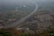 A long highway stretches across a lush, green landscape, elevated on rugged terrain. The road is surrounded by dense vegetation and hills, with scattered trees across the scene. Vehicles are seen traveling along the highway, emphasizing a sense of movement within the tranquil environment.