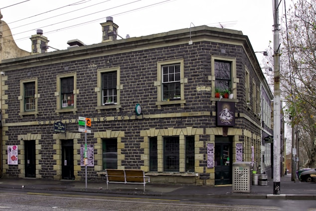 A two-story building constructed from dark stone with light stone trim, displaying signs for a hotel and bar. It features several windows and is located on a street corner with a bus stop and bench nearby. The street is lined with leafless trees and there are advertisements and signs visible.