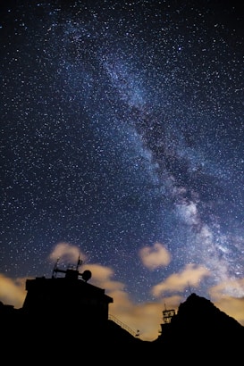 A breathtaking view of the Milky Way galaxy stretching across the night sky, with countless stars visible. Silhouettes of an observatory and mountains are seen in the foreground, adding an element of mystery and science. Fluffy clouds softly illuminated by the starlight accentuate the scene.