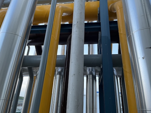 A series of industrial pipes and structures, featuring a mix of metallic and painted surfaces running vertically and horizontally. The pipes are primarily shiny and metallic, with some painted in bright yellow. The design is complex, with the arrangement creating a network of lines and shapes.