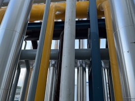 A series of industrial pipes and structures, featuring a mix of metallic and painted surfaces running vertically and horizontally. The pipes are primarily shiny and metallic, with some painted in bright yellow. The design is complex, with the arrangement creating a network of lines and shapes.