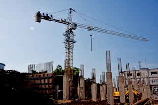 A construction site with a large crane towering above. Concrete columns are in various stages of completion, surrounded by rebar and construction materials. A bright sky forms the backdrop.