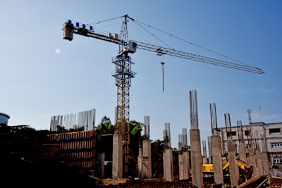 A construction site with a large crane towering above. Concrete columns are in various stages of completion, surrounded by rebar and construction materials. A bright sky forms the backdrop.
