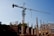 A construction site with a large crane towering above. Concrete columns are in various stages of completion, surrounded by rebar and construction materials. A bright sky forms the backdrop.