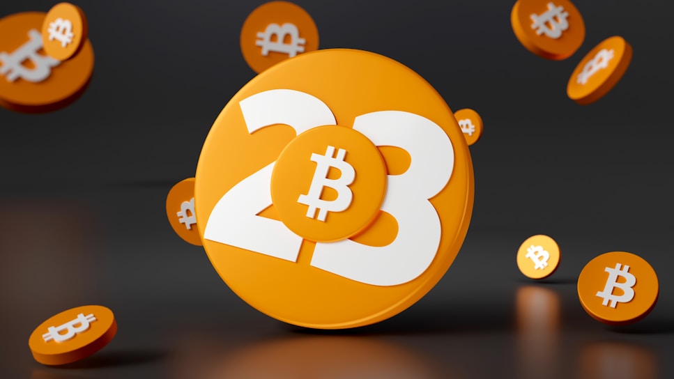 A large orange disc with the number 23 prominently displayed in white, accompanied by the Bitcoin symbol. Smaller orange discs with Bitcoin symbols surround the main disc, all set against a dark background.