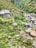 A lush, mountainous landscape featuring a small stone house with a blue roof and another structure with a thatched roof. Colorful prayer flags are strung along paths that meander through the green terrain. A rocky stream flows nearby, adding to the serene and natural setting.