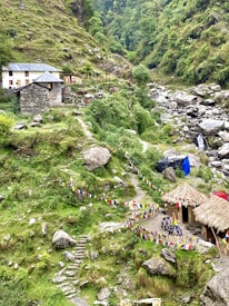 A lush, mountainous landscape featuring a small stone house with a blue roof and another structure with a thatched roof. Colorful prayer flags are strung along paths that meander through the green terrain. A rocky stream flows nearby, adding to the serene and natural setting.