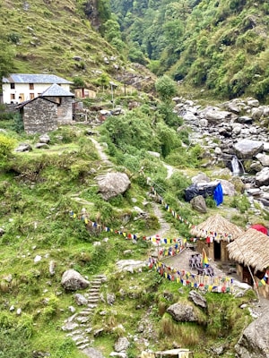 A lush, mountainous landscape featuring a small stone house with a blue roof and another structure with a thatched roof. Colorful prayer flags are strung along paths that meander through the green terrain. A rocky stream flows nearby, adding to the serene and natural setting.