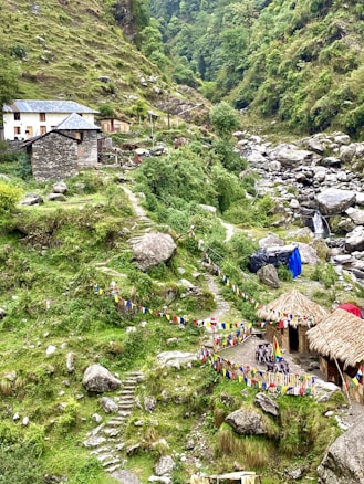 A lush, mountainous landscape featuring a small stone house with a blue roof and another structure with a thatched roof. Colorful prayer flags are strung along paths that meander through the green terrain. A rocky stream flows nearby, adding to the serene and natural setting.