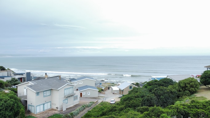 Several modern coastal houses with blue and gray tones are situated near a tranquil ocean. Rolling waves approach a rocky shoreline and a mix of lush green trees surround the area, creating a serene atmosphere.