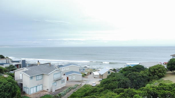 Several modern coastal houses with blue and gray tones are situated near a tranquil ocean. Rolling waves approach a rocky shoreline and a mix of lush green trees surround the area, creating a serene atmosphere.