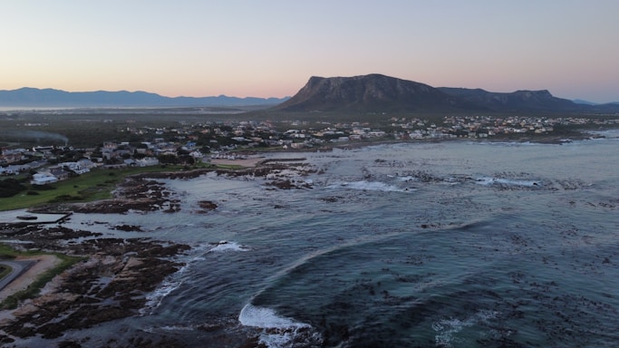 A coastal landscape with an expansive view of the ocean and rocky shoreline in the foreground. Numerous houses are scattered across the green land behind the shoreline. The scene is framed by a dramatic mountain range in the distance, under a clear sky transitioning from day to dusk.