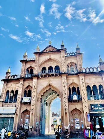 A grand historic gateway with intricate architectural details and arches stands tall against a vibrant blue sky. The structure is adorned with multiple tiers, pointed minarets, and decorative patterns. People and vehicles are seen passing through the gateway, adding a lively urban atmosphere.